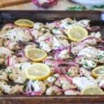 Side view of sheet pan Mediterranean chicken with red onions, lemons, and dollops of tzatziki.