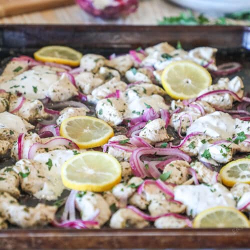 Side view of sheet pan Mediterranean chicken with red onions, lemons, and dollops of tzatziki.