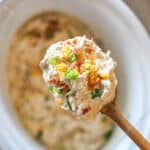 Crockpot Million Dollar Chicken- in a white crockpot- being lifted out by wooden spoon
