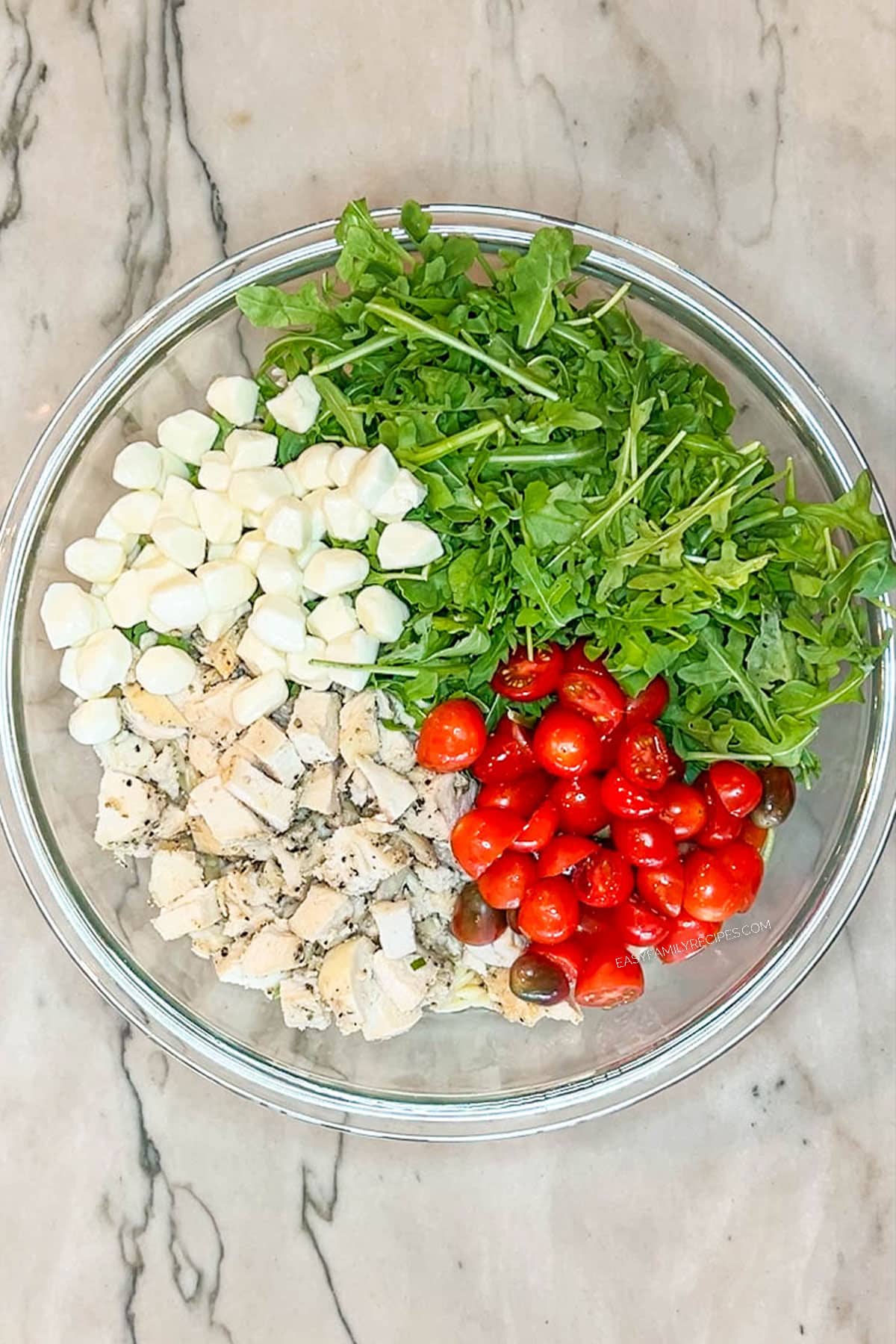 arugula, tomatoes, mozzarella pearls and chopped chicken in glass bowl- on a marble background