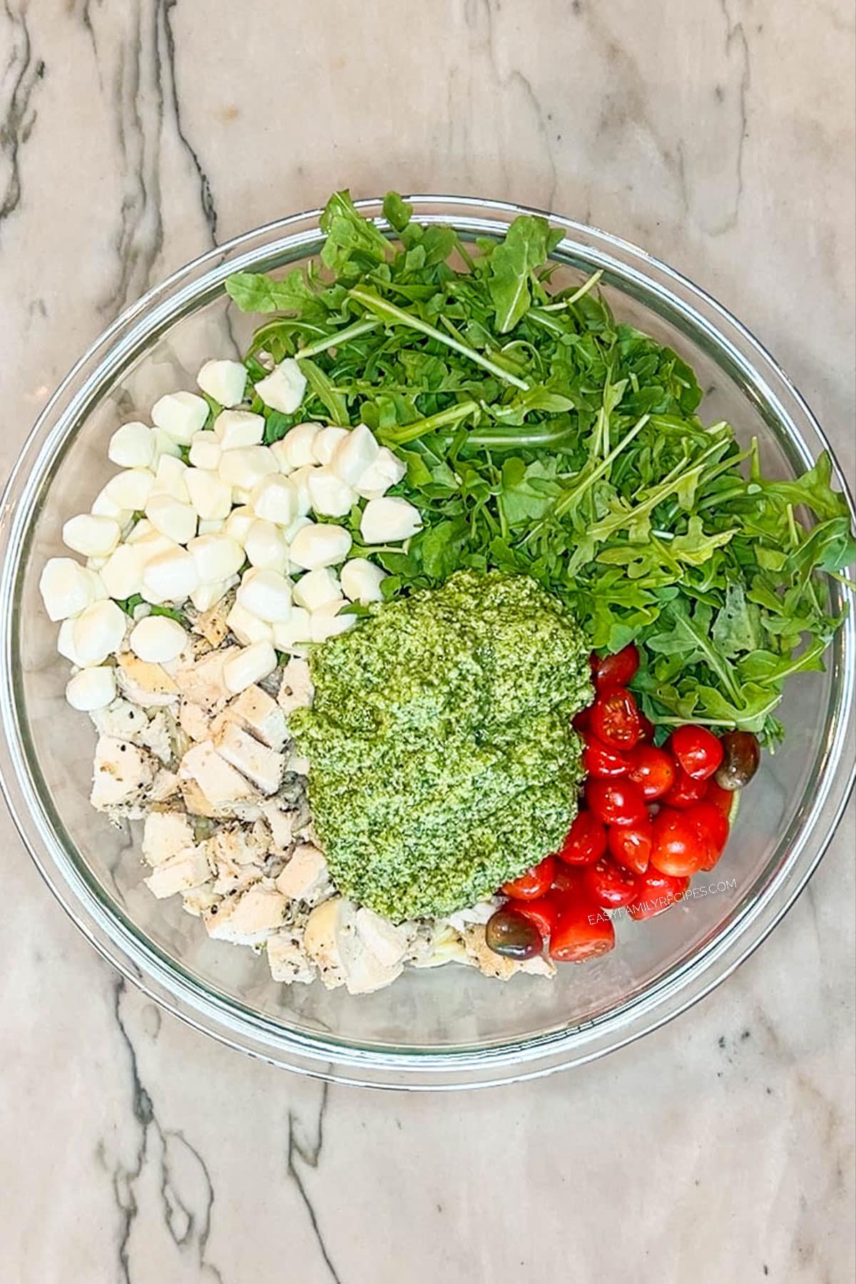 arugula, tomatoes, mozzarella pearls, chopped chicken and pesto in glass bowl- on a marble background