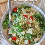 Pesto chicken salad with orzo in white bowl with fork. On a wooden backdrop with a kitchen towel and wooden, cutting board in background