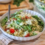 Pesto chicken salad with orzo in white bowl with fork. On a wooden backdrop with a kitchen towel and wooden, cutting board in background