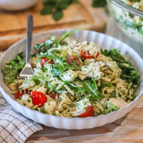 Pesto chicken salad with orzo in white bowl with fork. On a wooden backdrop with a kitchen towel and wooden, cutting board in background