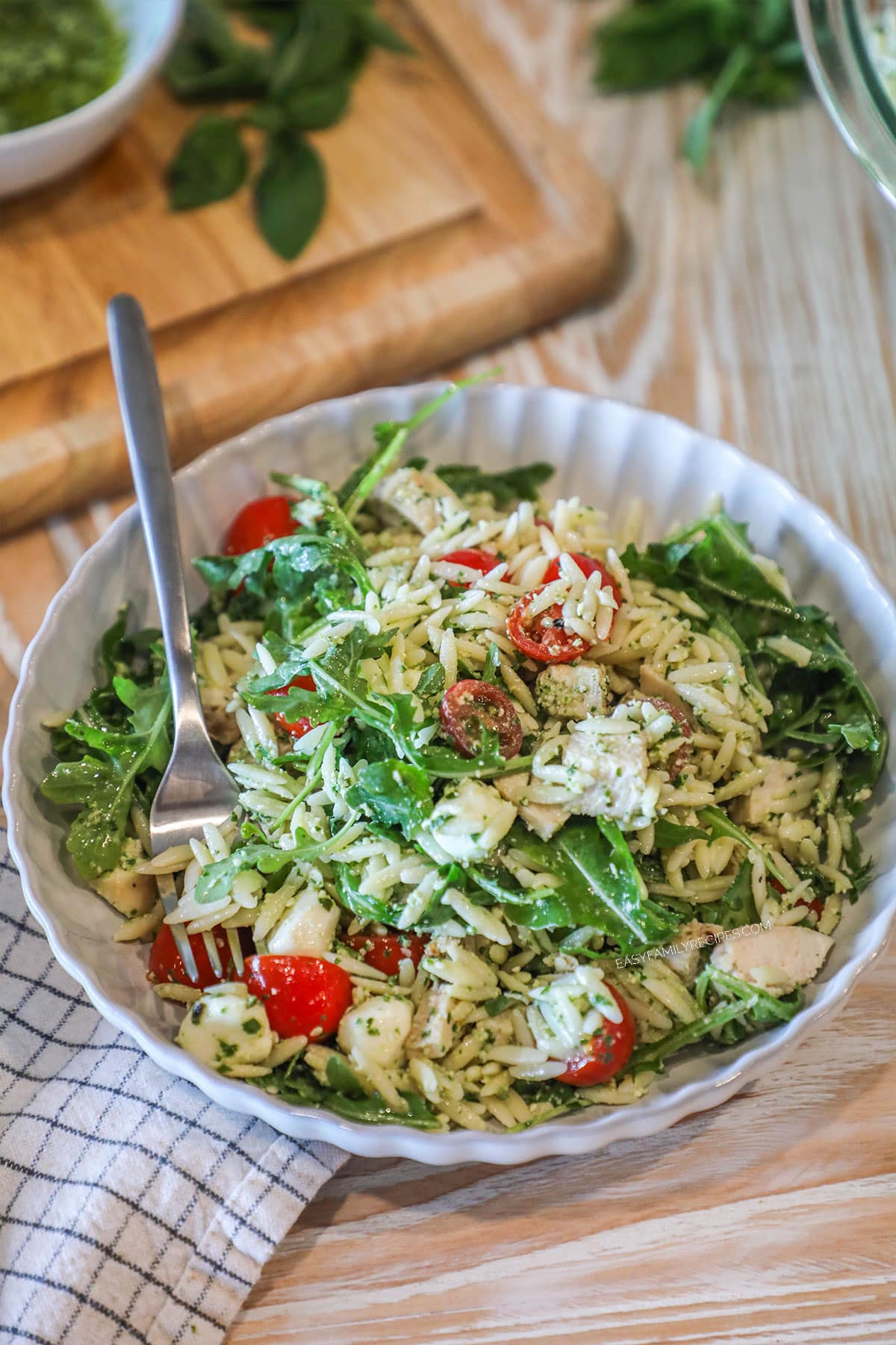 Pesto chicken salad with fork in white bowl with spoon. On a wooden backdrop with a kitchen towel and wooden, cutting board in background
