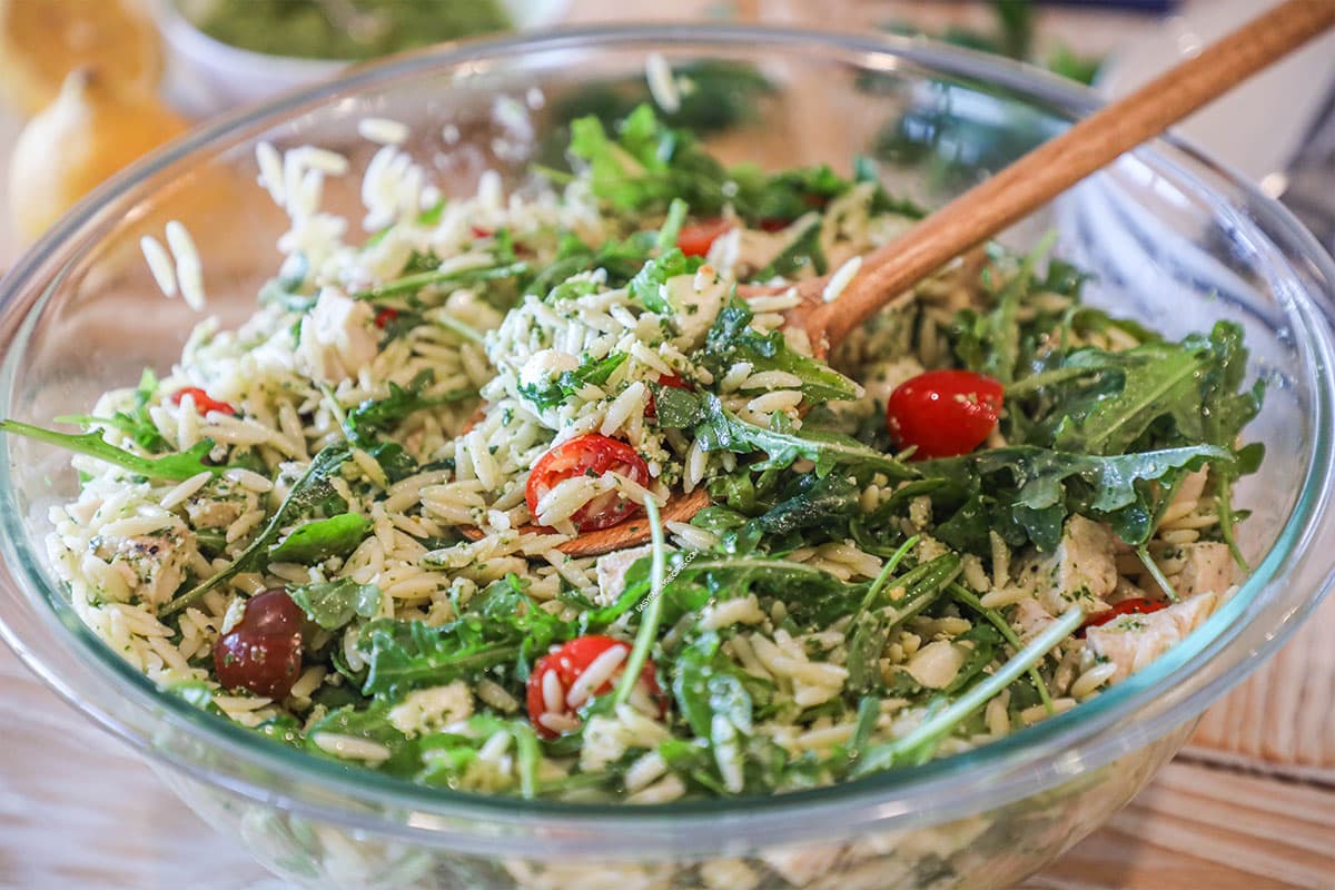 Pesto chicken salad with orzo in large, glass bowl with wooden spoon