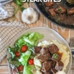 balsamic steak bites over mashed potatoes with a side salad- on a white plate- pan of steak bites, head of garlic and towel in the background