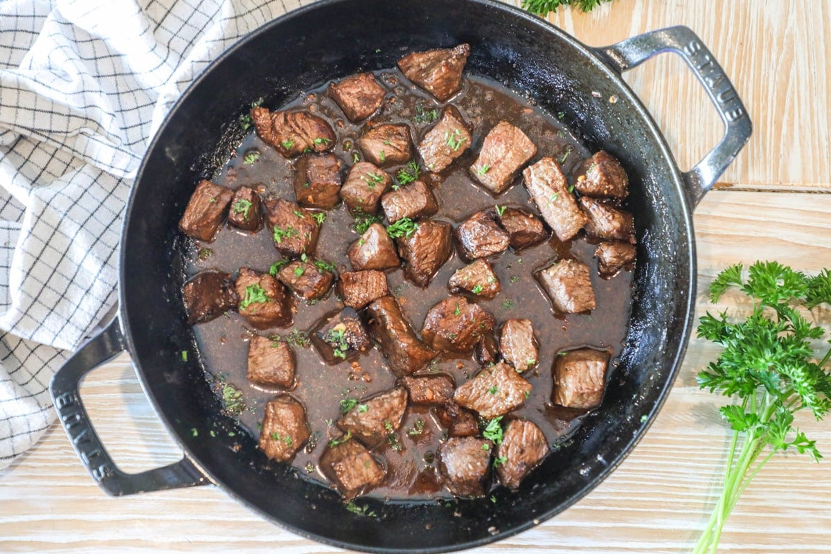 balsamic steak bites seared and cooked in cast iron skillet- on a wooden background with a towel and fresh herbs