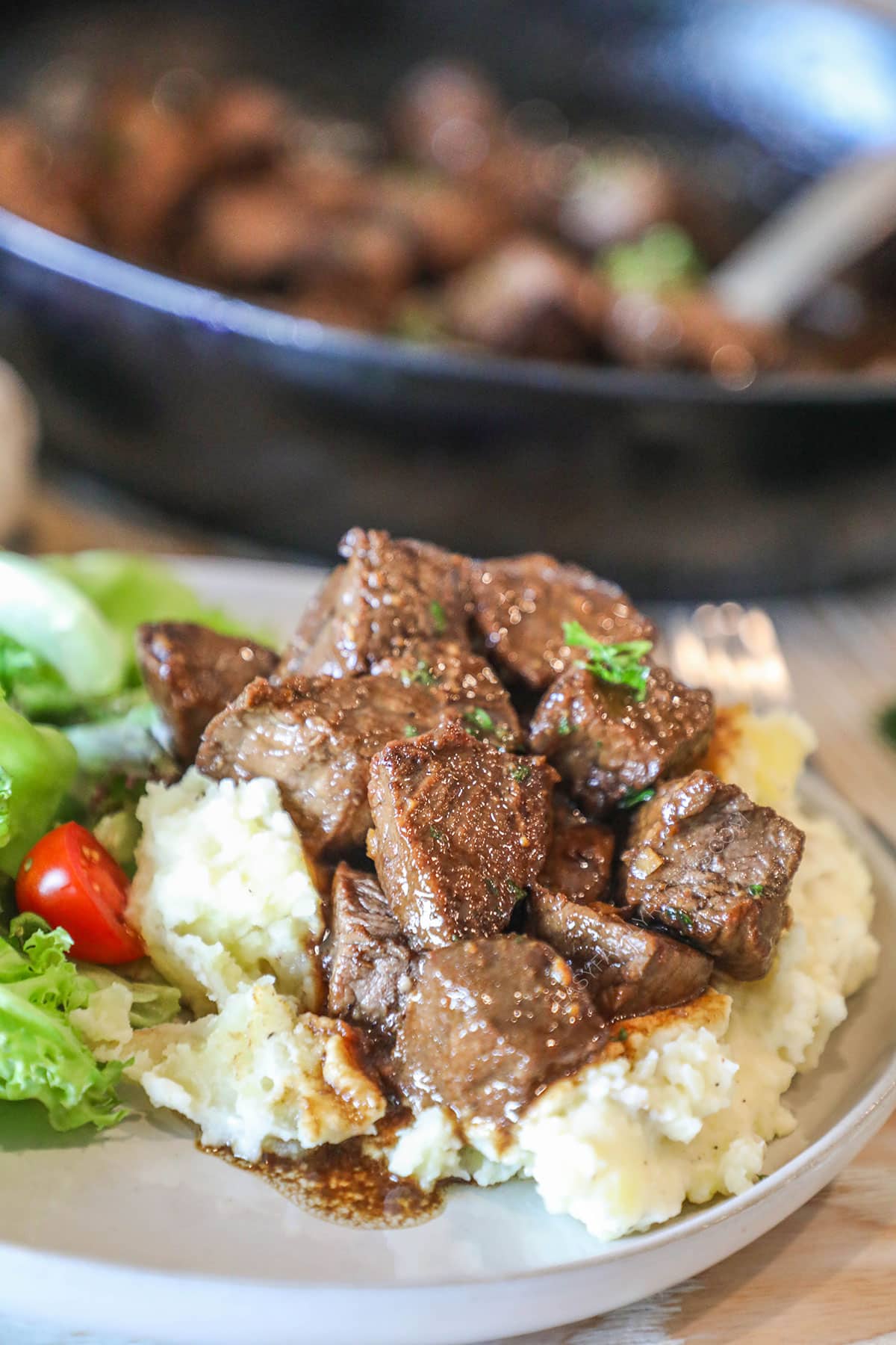 balsamic steak bites over mashed potatoes with a side salad- on a white plate- pan of steak bites in the background