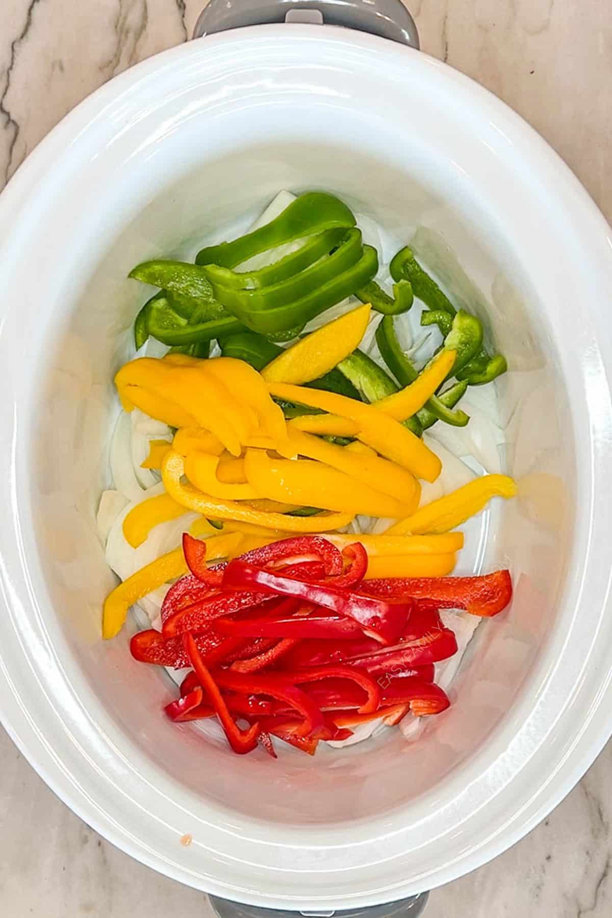 Peppers and onions layered in the bottom of a slow cooker.