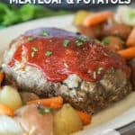 Crockpot meatloaf plated on a white platter alongside baby carrots and new potatoes. On a wooden table with a grey crockpot in the background.