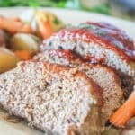 Crockpot meatloaf sliced on a white platter- with baby carrots and new potatoes