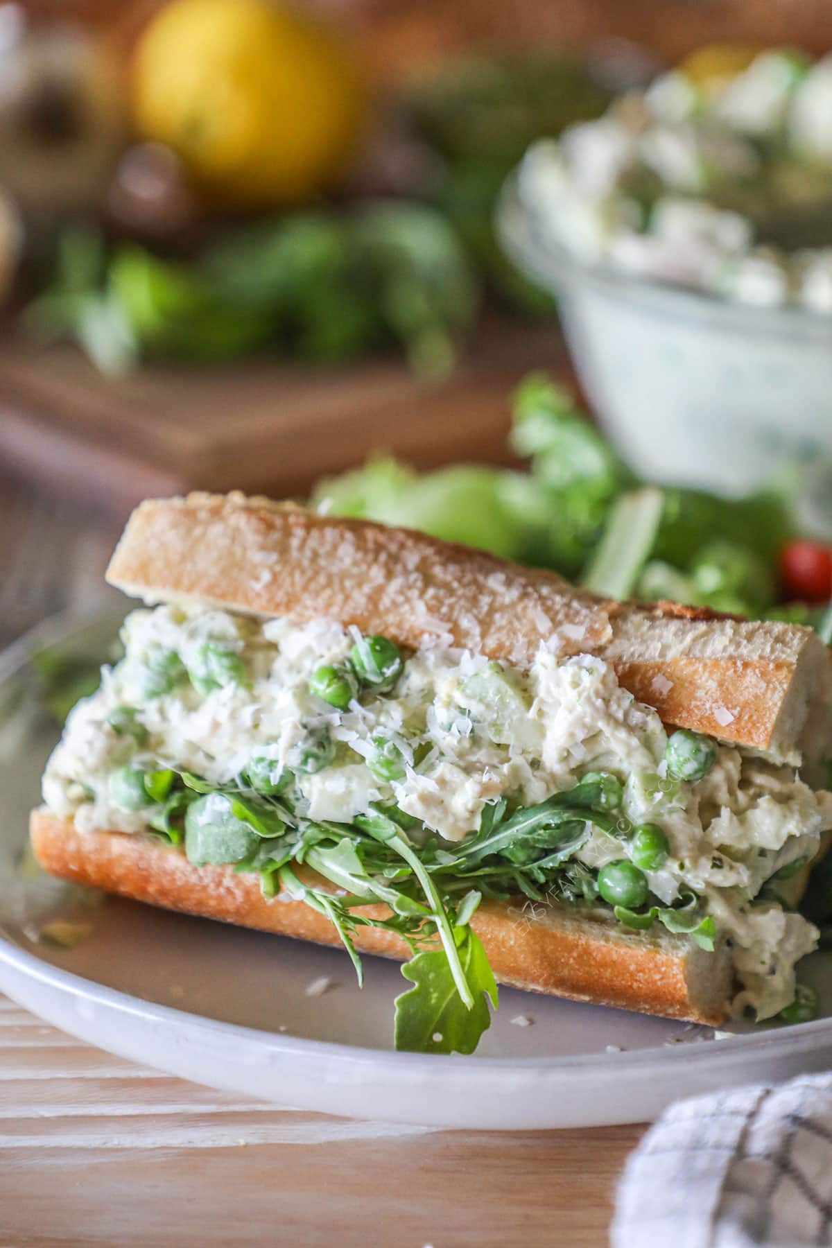 Baguette stuffed with arugula and pesto chicken salad- on a white plate. Wooden board, lemon and bowl of chicken salad in background