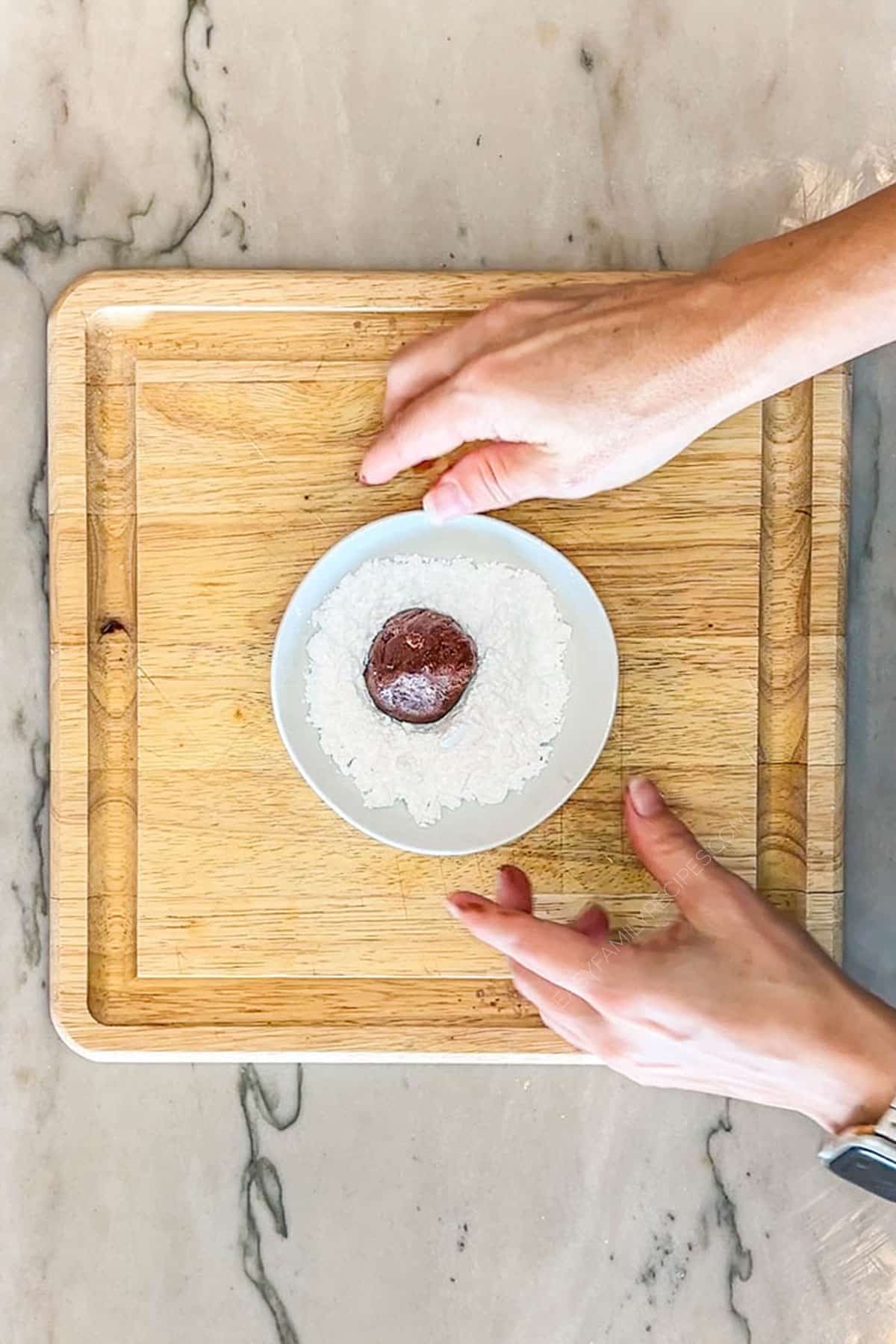 Step 3 for making Brownie Crinkle Cookies- roll cookie dough ball in bowl of powdered sugar- image includes white bowl on a wooden board, with two hands