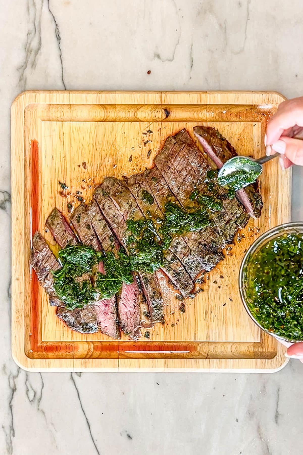Sliced flank steak topped with chimichurri sauce- on a wooden cutting board with a bowl of chimichurri on the side