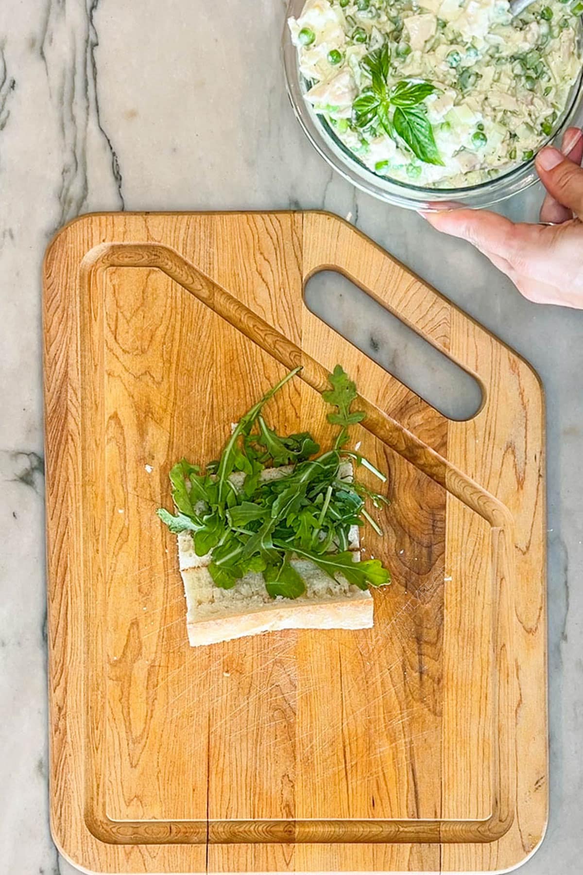 Step three for making pesto chicken salad- slice open a baguette and lay fresh arugula. On a wooden board with bowl of chicken salad in background