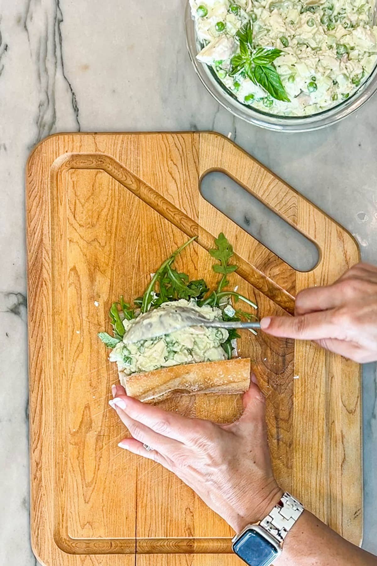Step four for making pesto chicken salad- add spoonful of chicken salad to baguette. On a wooden board with bowl of chicken salad in background