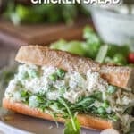 Baguette stuffed with arugula and pesto chicken salad- on a white plate. Wooden board, lemon and bowl of chicken salad in background