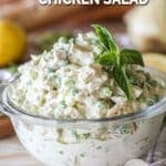 Glass bowl with pesto chicken salad and fresh basil leaves on top. On a wooden background with a baguette and lemon in background.
