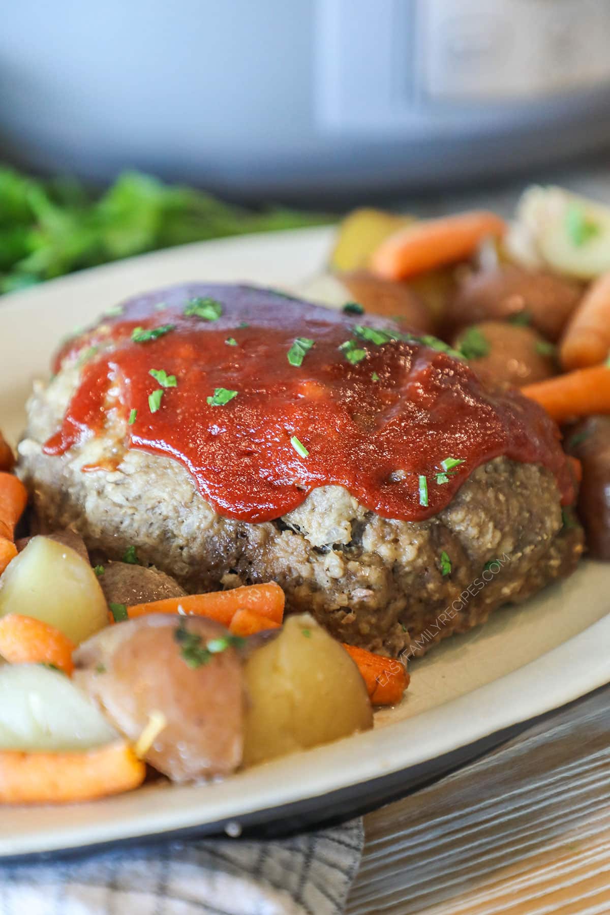 Crockpot meatloaf plated on a white platter alongside baby carrots and new potatoes. On a wooden table with a grey crockpot in the background.