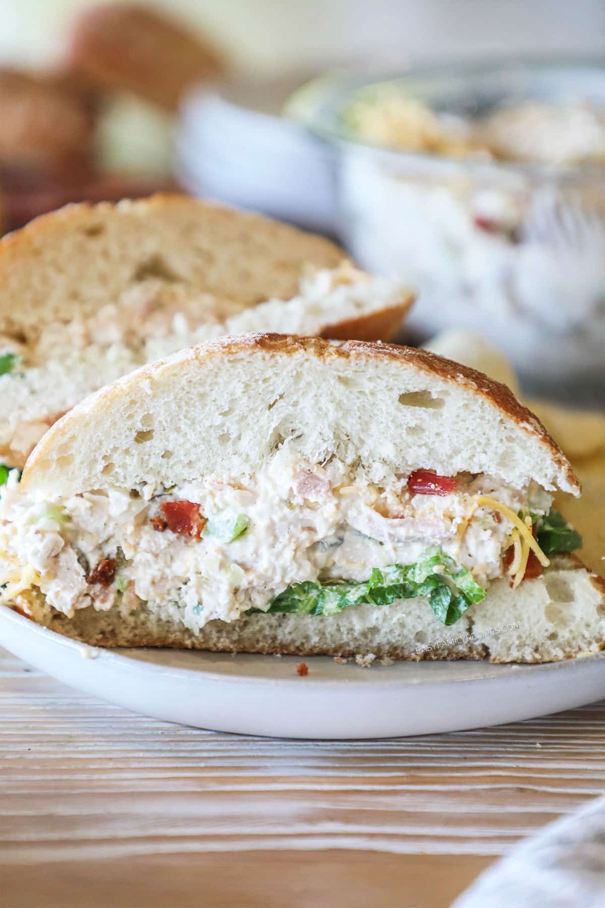 Jalapeño Popper Chicken Salad sandwich, cut in half, on a white dish. On a wooden background with a bowl of chicken salad.