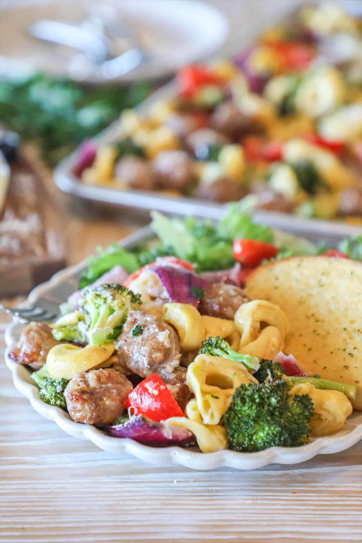 Individual serving of Sheet Pan Italian Sausage and Cheese Tortellini on a white plate with side salad and garlic bread. Full sheet pan is in background