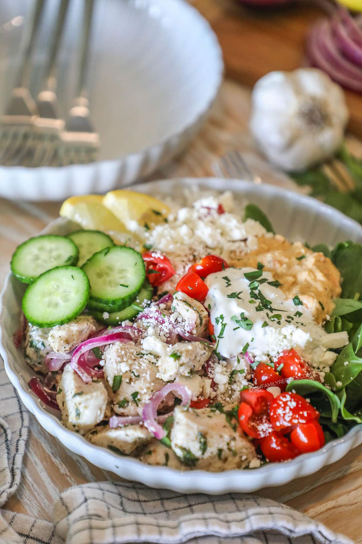 Completed Greek Chicken Bowl in a white bowl, on a wooden background, with a kitchen towel, extra white bowl and forks in background, and a head of garlic. Bowl contains rice, spinach, chicken, cucumbers, tomatoes, tzatziki, hummus, feta and lemon slice