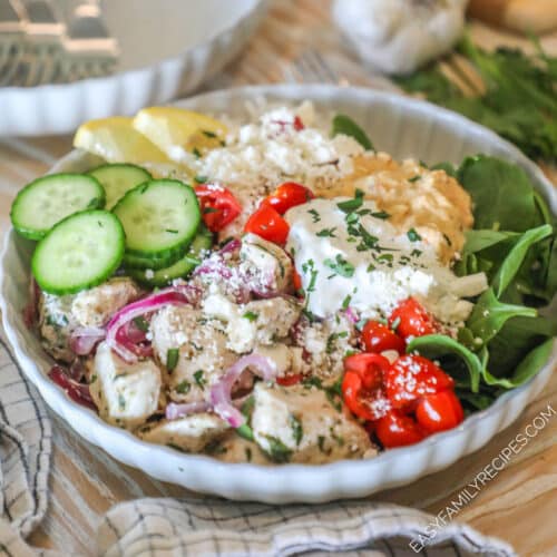 Completed Greek Chicken Bowl in a white bowl, on a wooden background, with a kitchen towel, extra white bowl and forks in background, and a head of garlic. Bowl contains rice, spinach, chicken, cucumbers, tomatoes, tzatziki, hummus, feta and lemon slice