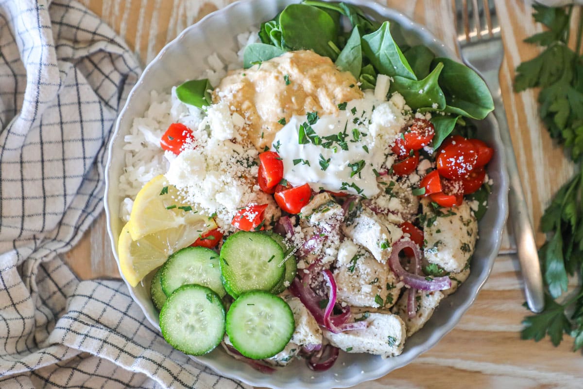 Completed Greek Chicken Bowl in a white bowl, on a wooden background, with a kitchen towel. Bowl contains rice, spinach, chicken, cucumbers, tomatoes, tzatziki, hummus, feta and lemon slice