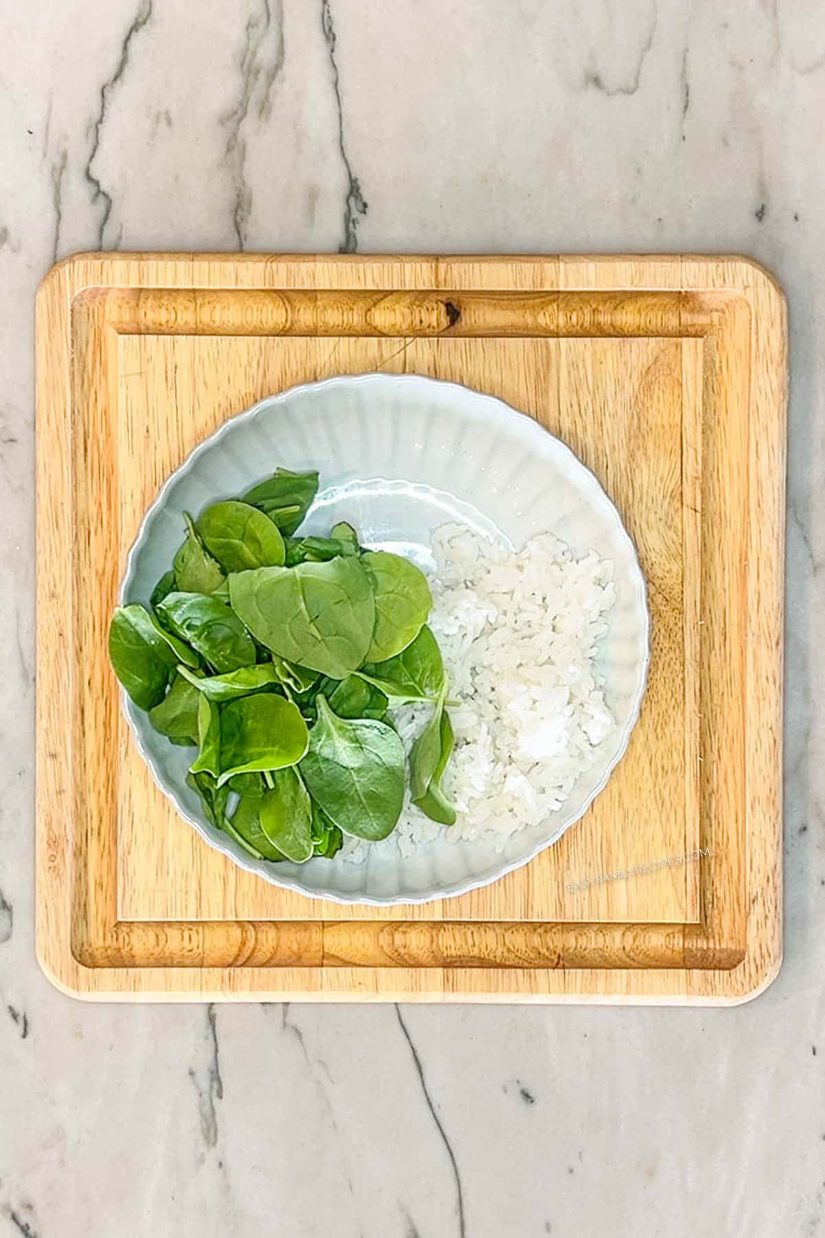 Step 2 for making Greek Chicken Bowls. White bowl with rice and fresh baby spinach. On a square wooden board on a marble background