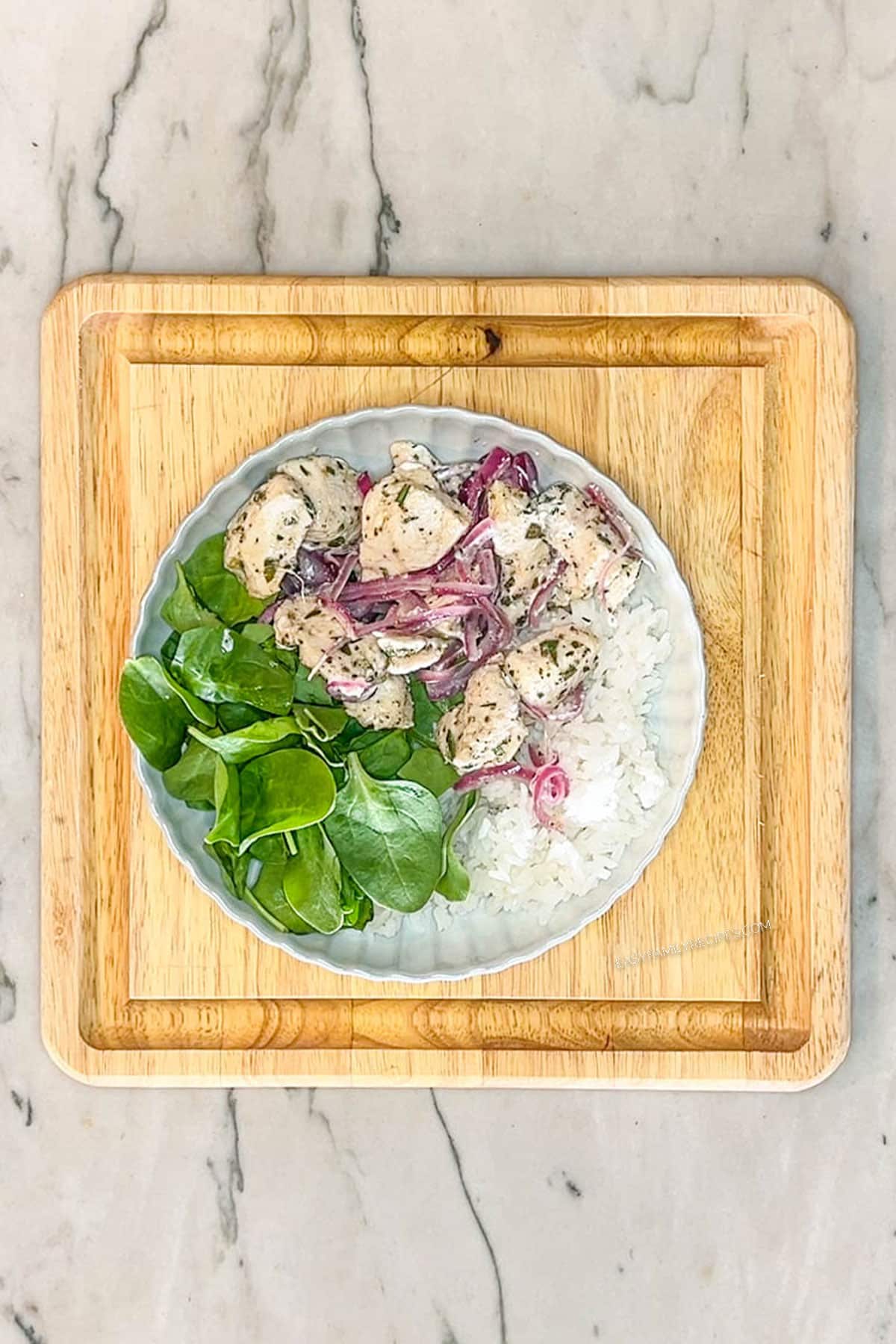 Step 3 for making Greek Chicken Bowls. White bowl with rice, fresh baby spinach and greek chicken and red onions. On a square wooden board on a marble background