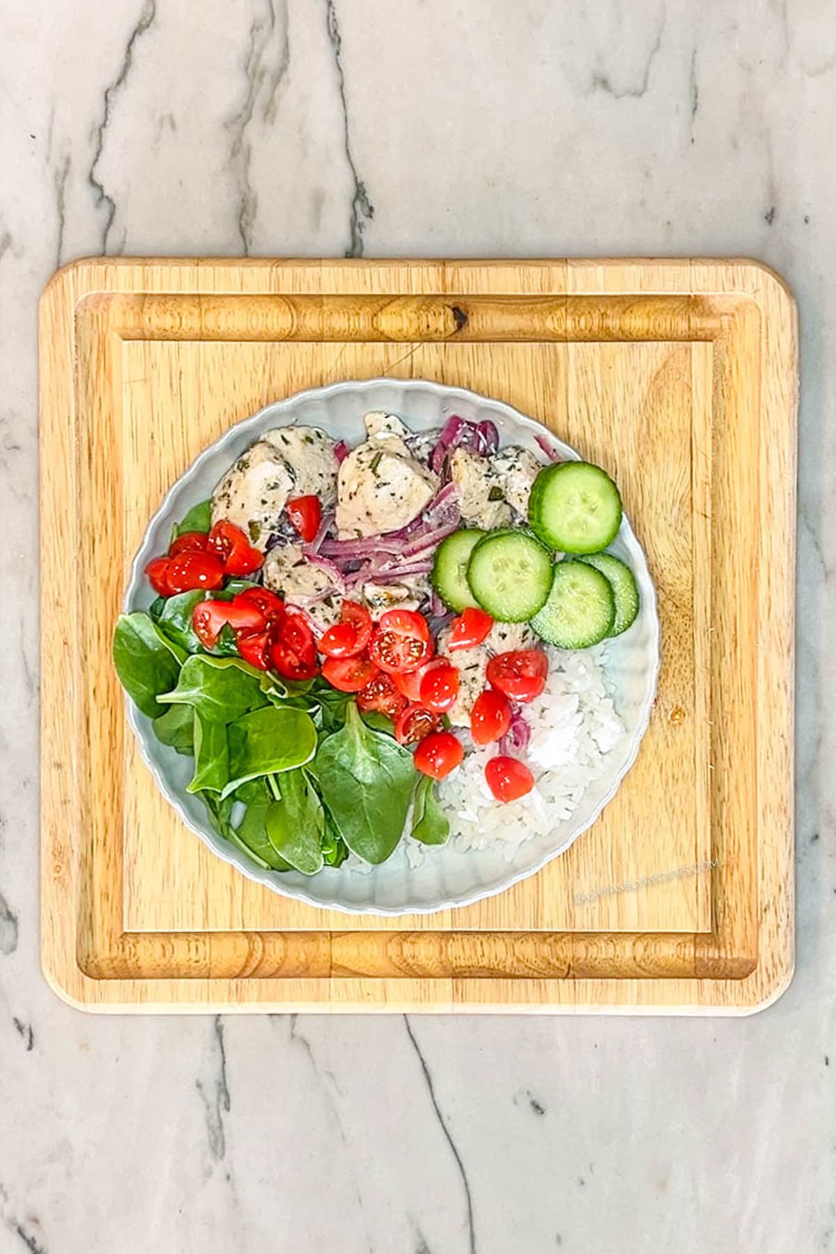 Step 4 for making Greek Chicken Bowls. White bowl with rice, fresh baby spinach, greek chicken and red onions, cucumbers and tomatoes. On a square wooden board on a marble background