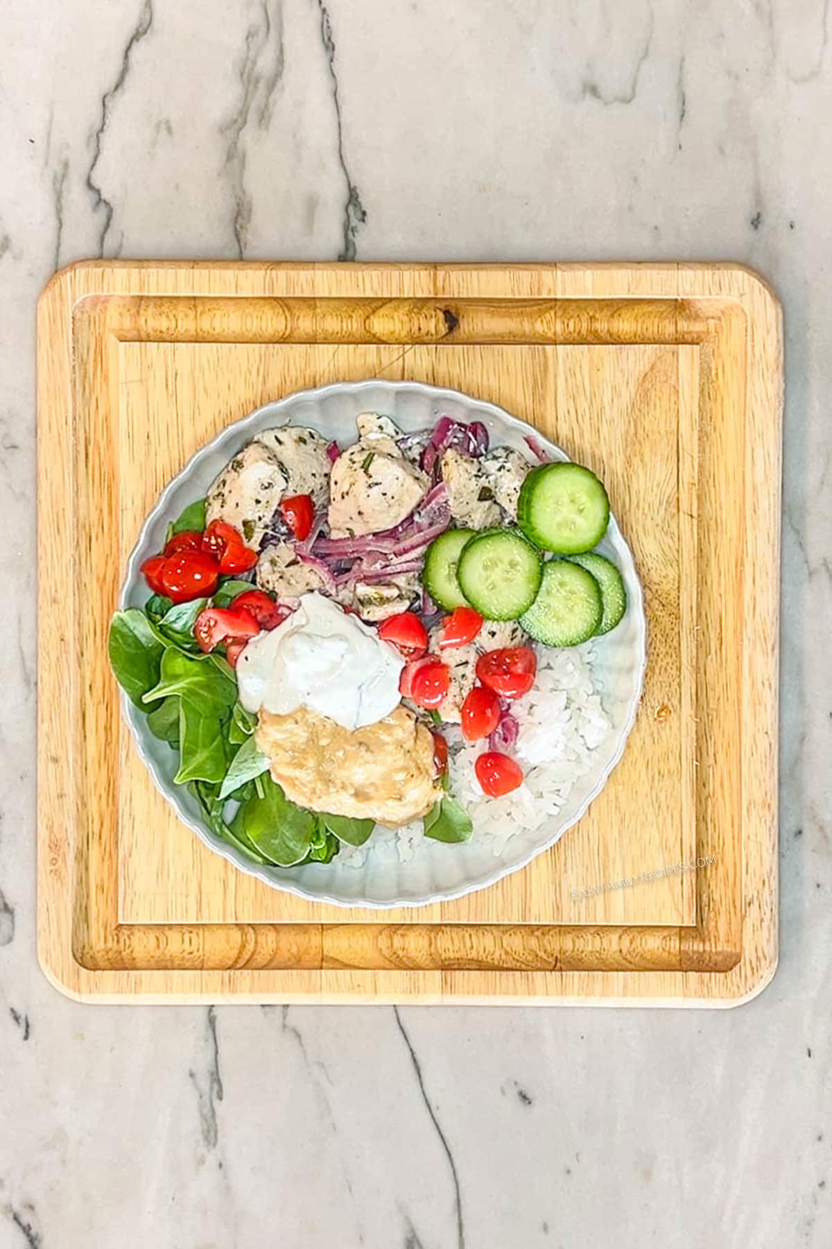 Step 5 for making Greek Chicken Bowls. White bowl with rice, fresh baby spinach, greek chicken and red onions, cucumbers, tomatoes feta, tzatziki and hummus. On a square wooden board on a marble background