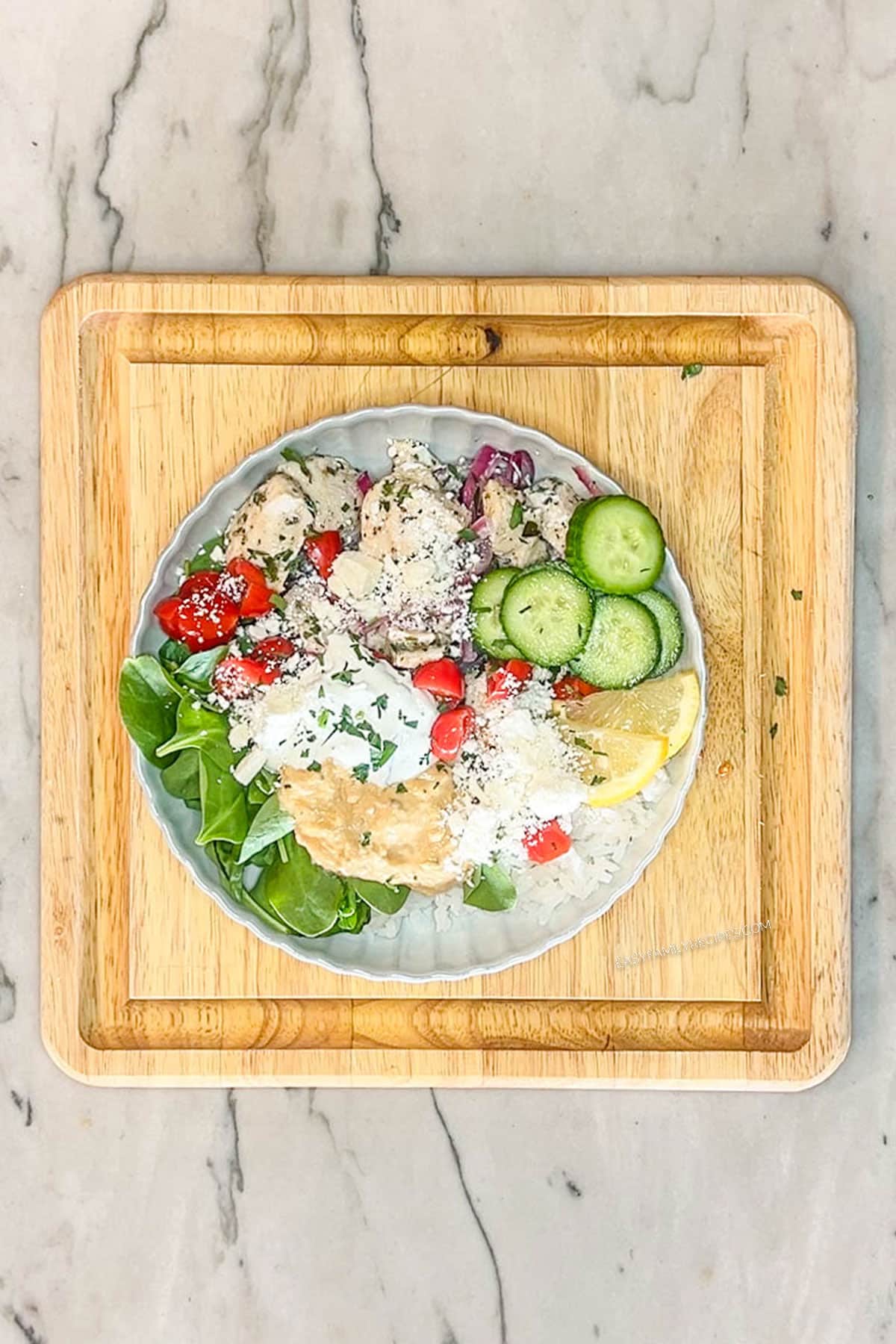 Completed Greek Chicken Bowl in a white bowl, on a square wooden board. Bowl contains rice, spinach, chicken, cucumbers, tomatoes, tzatziki, hummus, feta and lemon slice