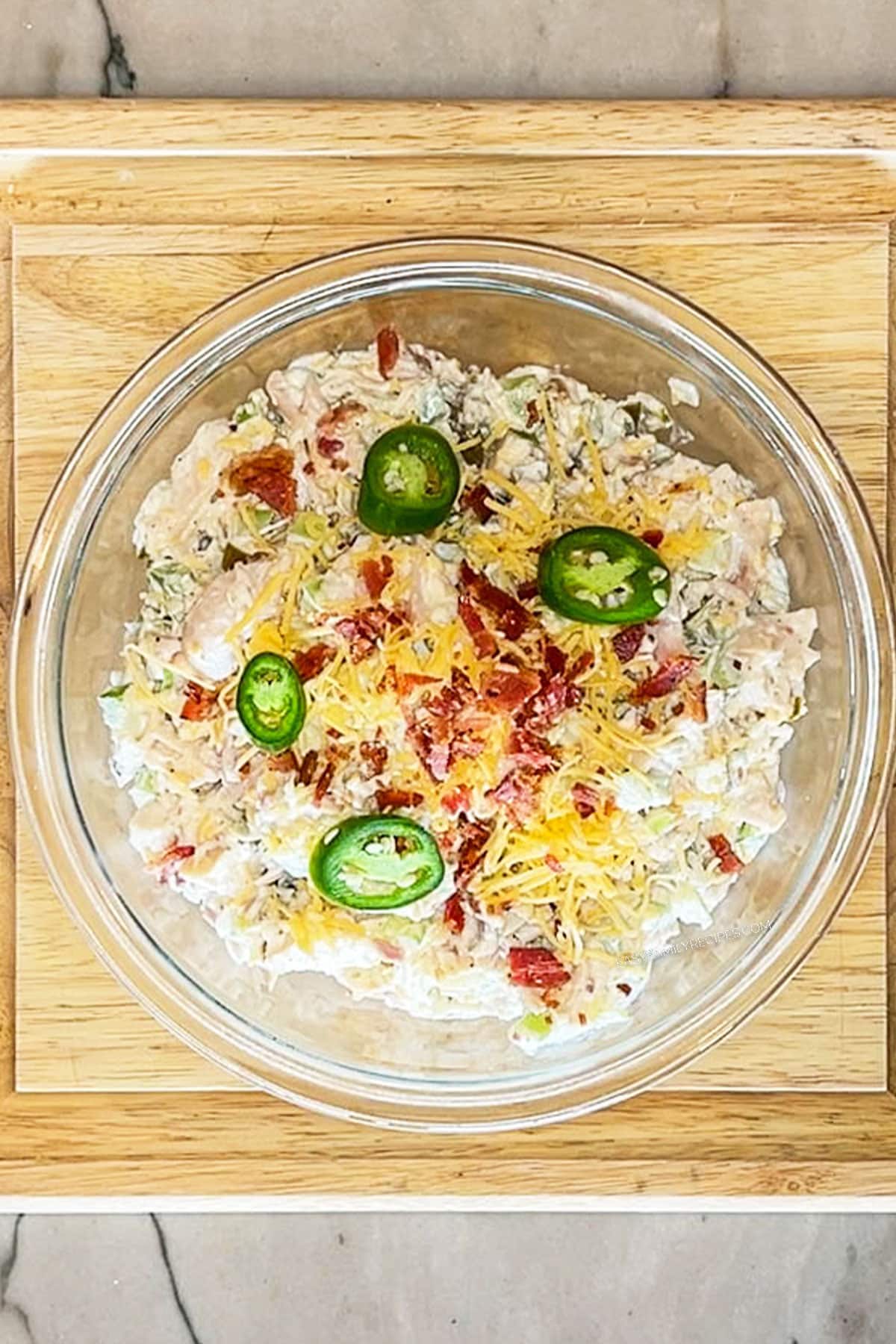 Step 4 for making Jalapeño Popper Chicken Salad. Combine all ingredients then top with cheddar, bacon and jalapeño slices. In a glass bowl on a wooden board.
