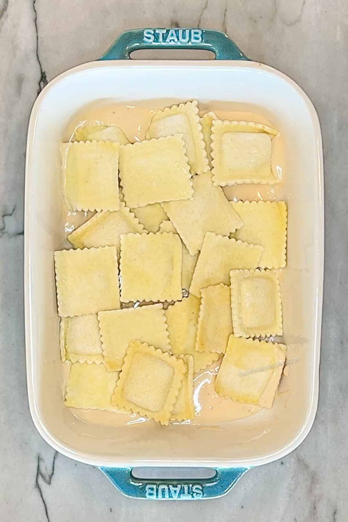 Step 1 for making Marry Me Baked Ravioli Casserole. Alfredo and ravioli in blue baking dish on a marble background