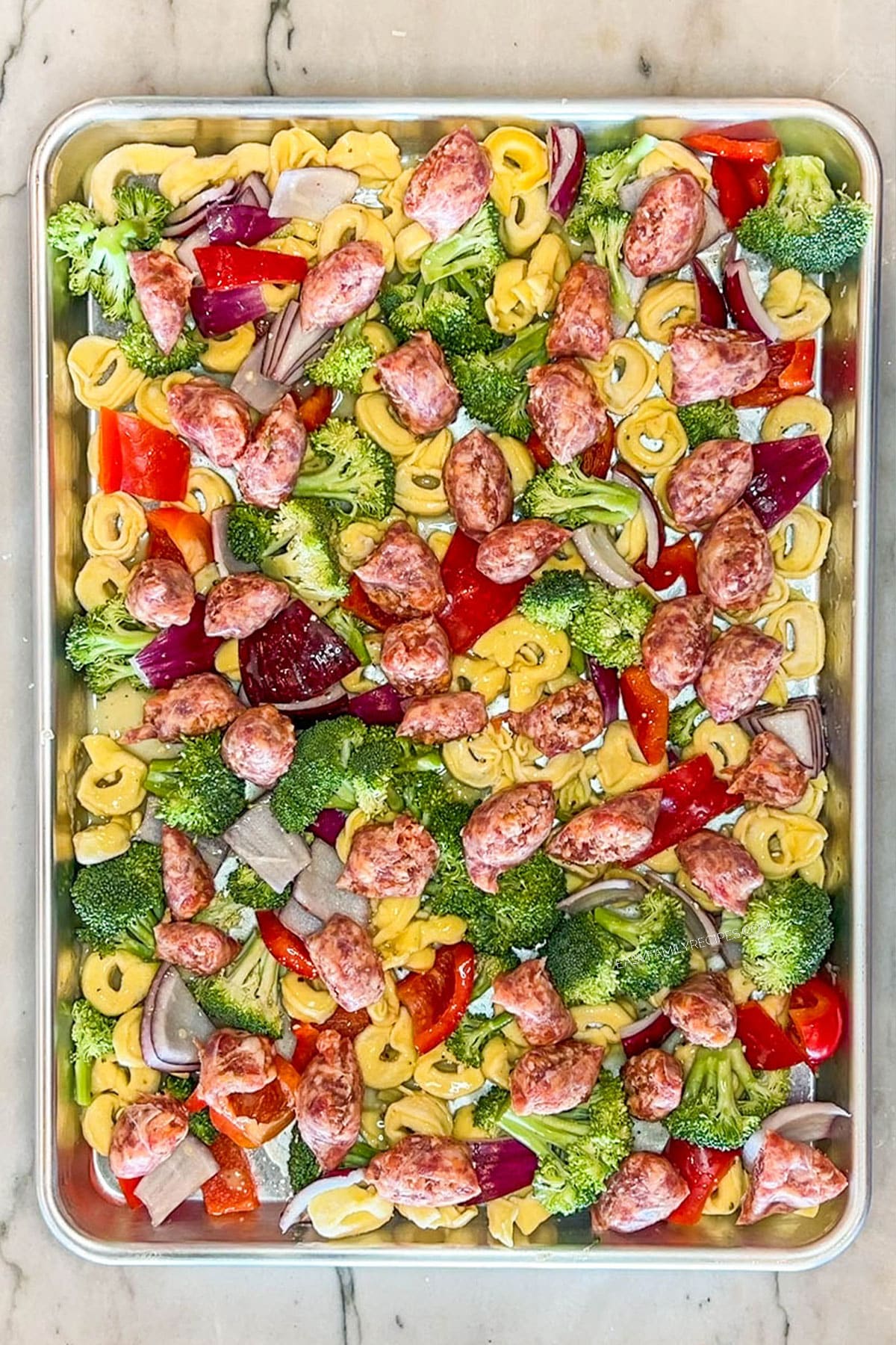 Step 3 to make Sheet Pan Italian Sausage Tortellini- Broccoli, tortellini, red peppers, red onions and Italian sausage on baking pan