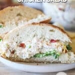 Jalapeño Popper Chicken Salad sandwich, cut in half, on a white dish. On a wooden background with a bowl of chicken salad and block of cheese behind.