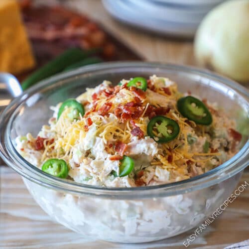 Glass bowl of Jalapeño Popper Chicken Salad on a wooden background with a block of cheddar cheese and onion.