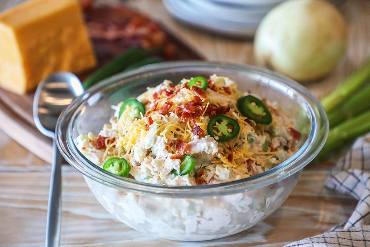 Glass bowl of Jalapeño Popper Chicken Salad on a wooden background with a block of cheddar cheese and onion.
