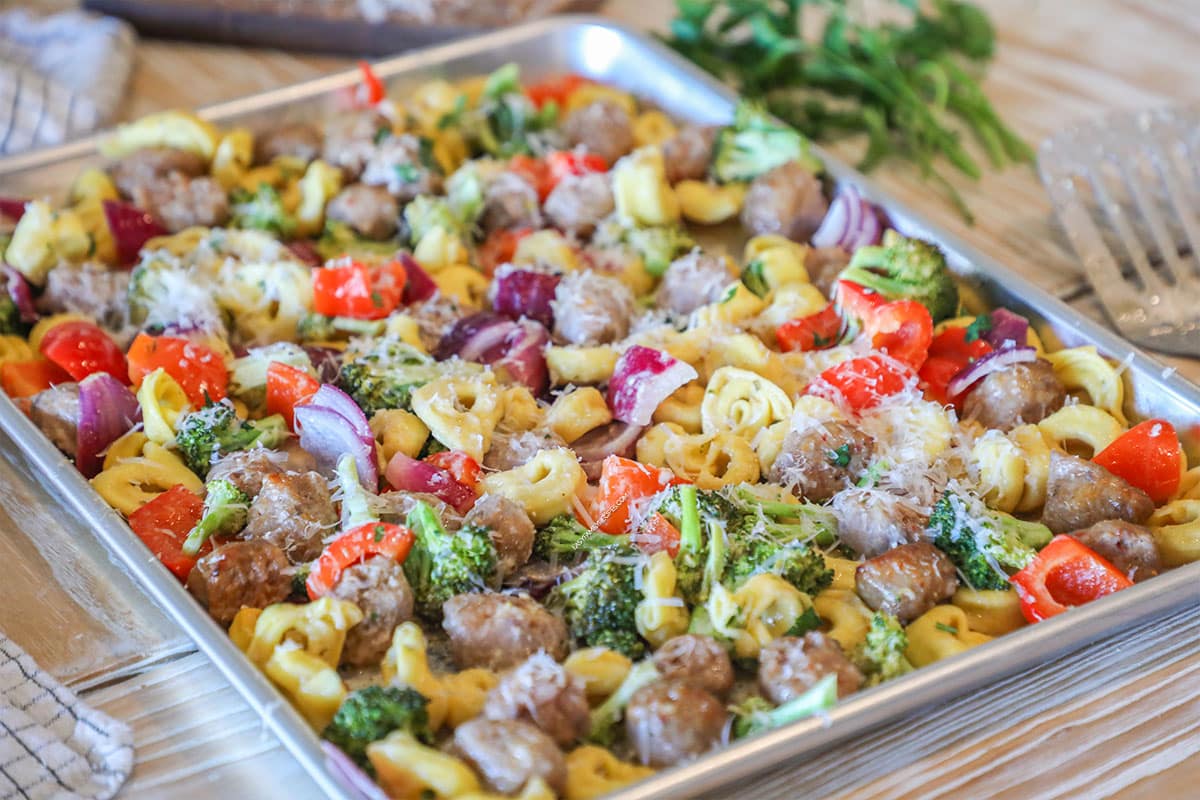 Sheet Pan Italian Sausage and Cheese Tortellini on a wooden background with fresh herbs and spatula