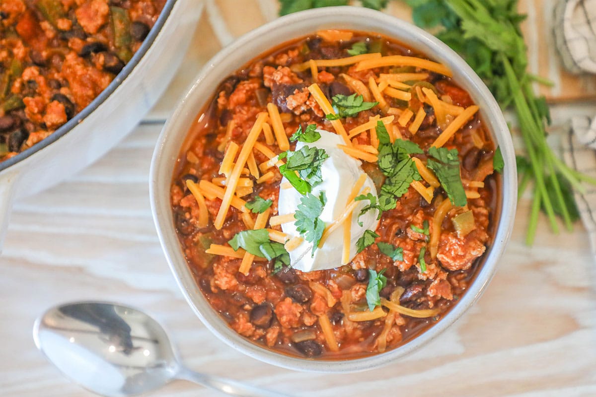 Individual serving of Chipotle Black Bean Ground Turkey Chili in a white bowl. On a wooden background with fresh herbs, a spoon and part of a large, white pot showing