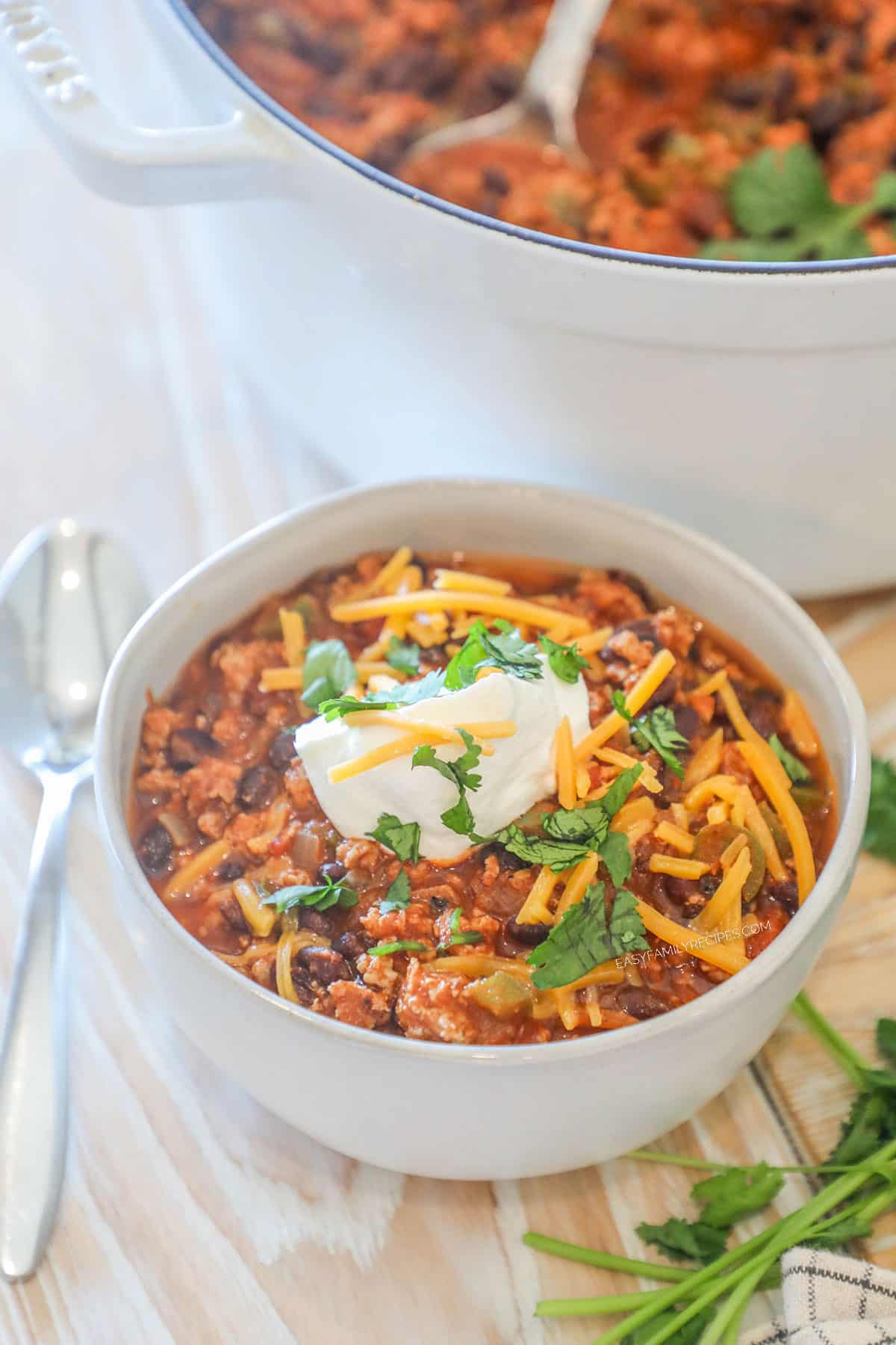 Individual serving of Chipotle Black Bean Ground Turkey Chili in a white bowl. On a wooden background with fresh herbs, a spoon and a large, white pot of chili