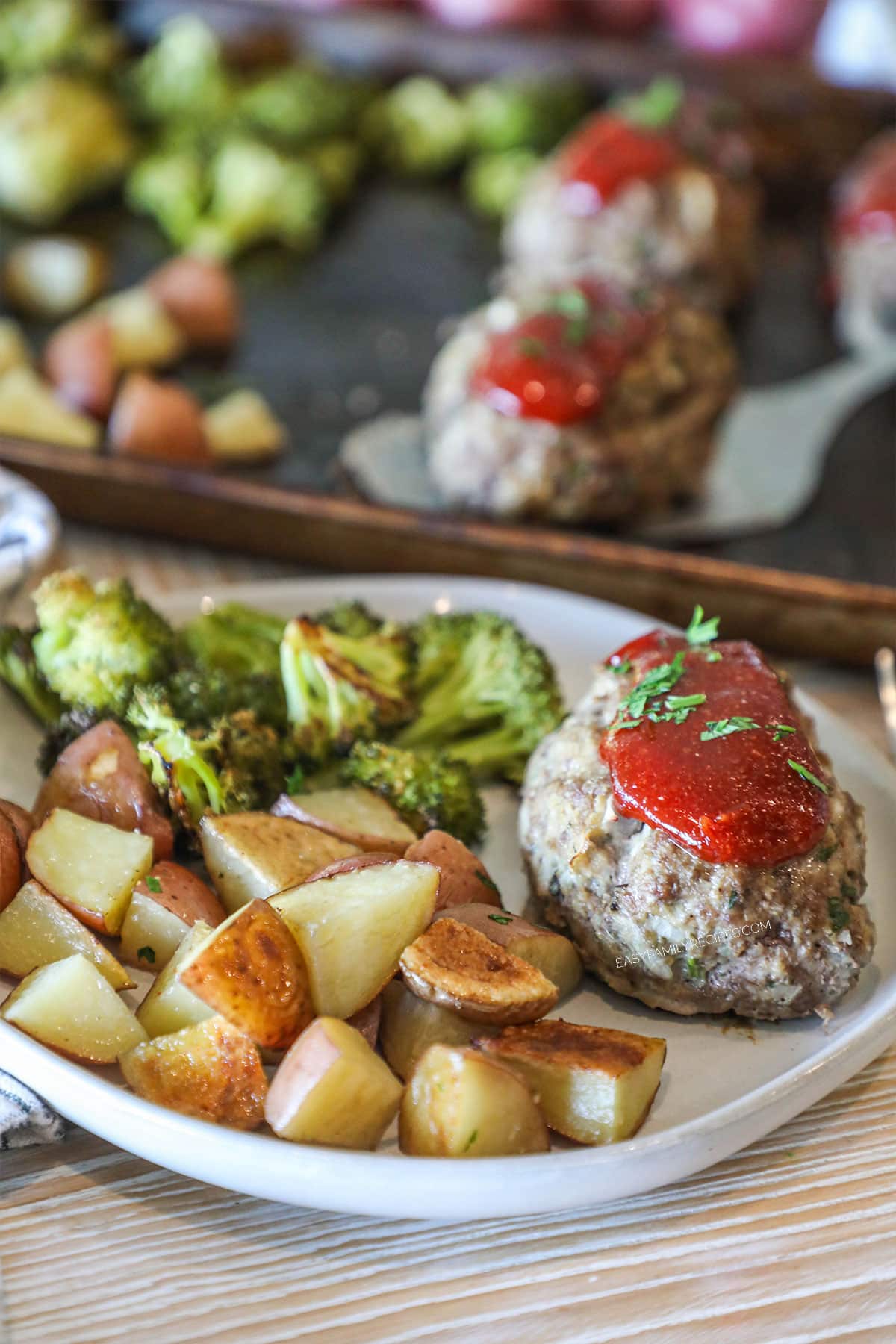 White plate with meatloaf, potatoes and broccoli. Pan of Mini Meatloaf Sheet Pan Dinner in background. On a wooden background