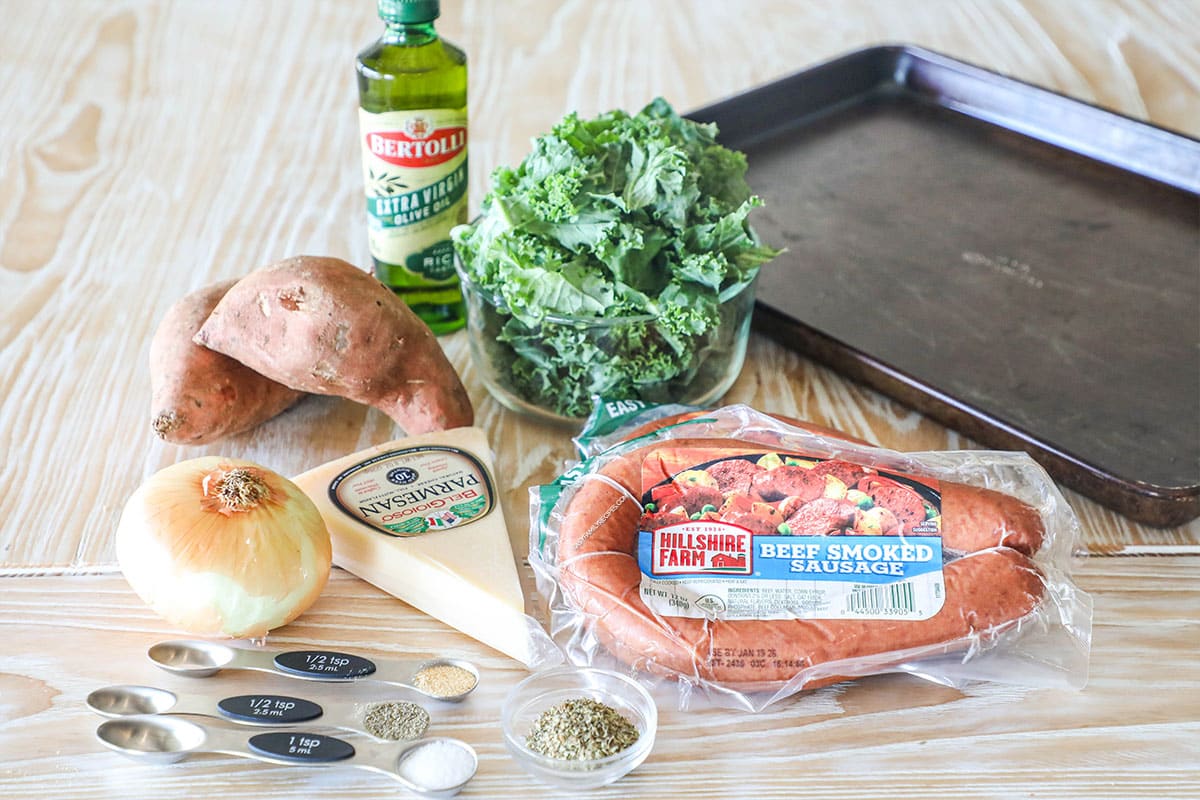 Ingredients for sheet pan sausage and sweet potatoes, including kale, smoked sausage, parmesan, oil, sweet potatoes, onions, and seasonings.