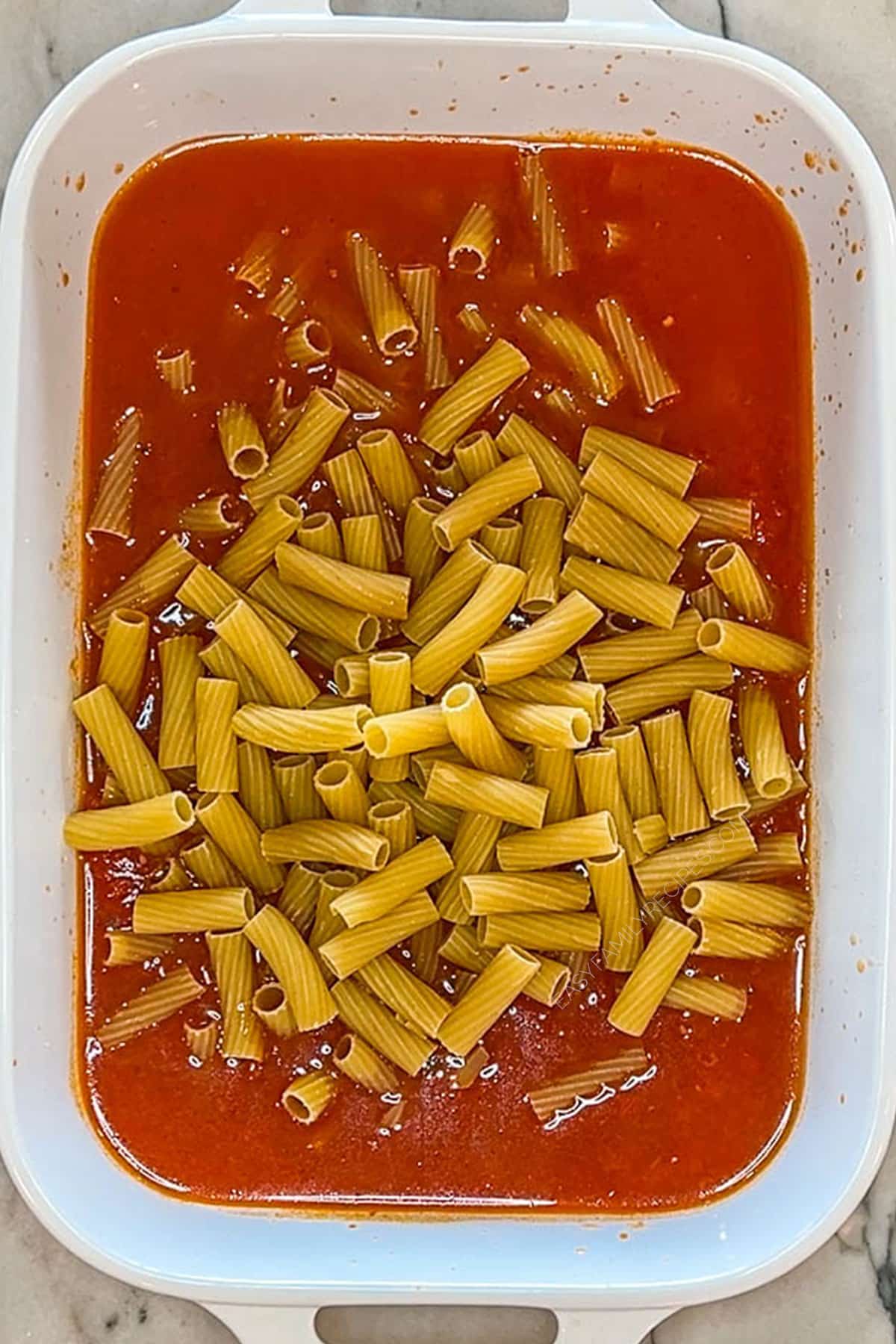 Dried pasta mixed with tomato sauce and broth in a baking dish.