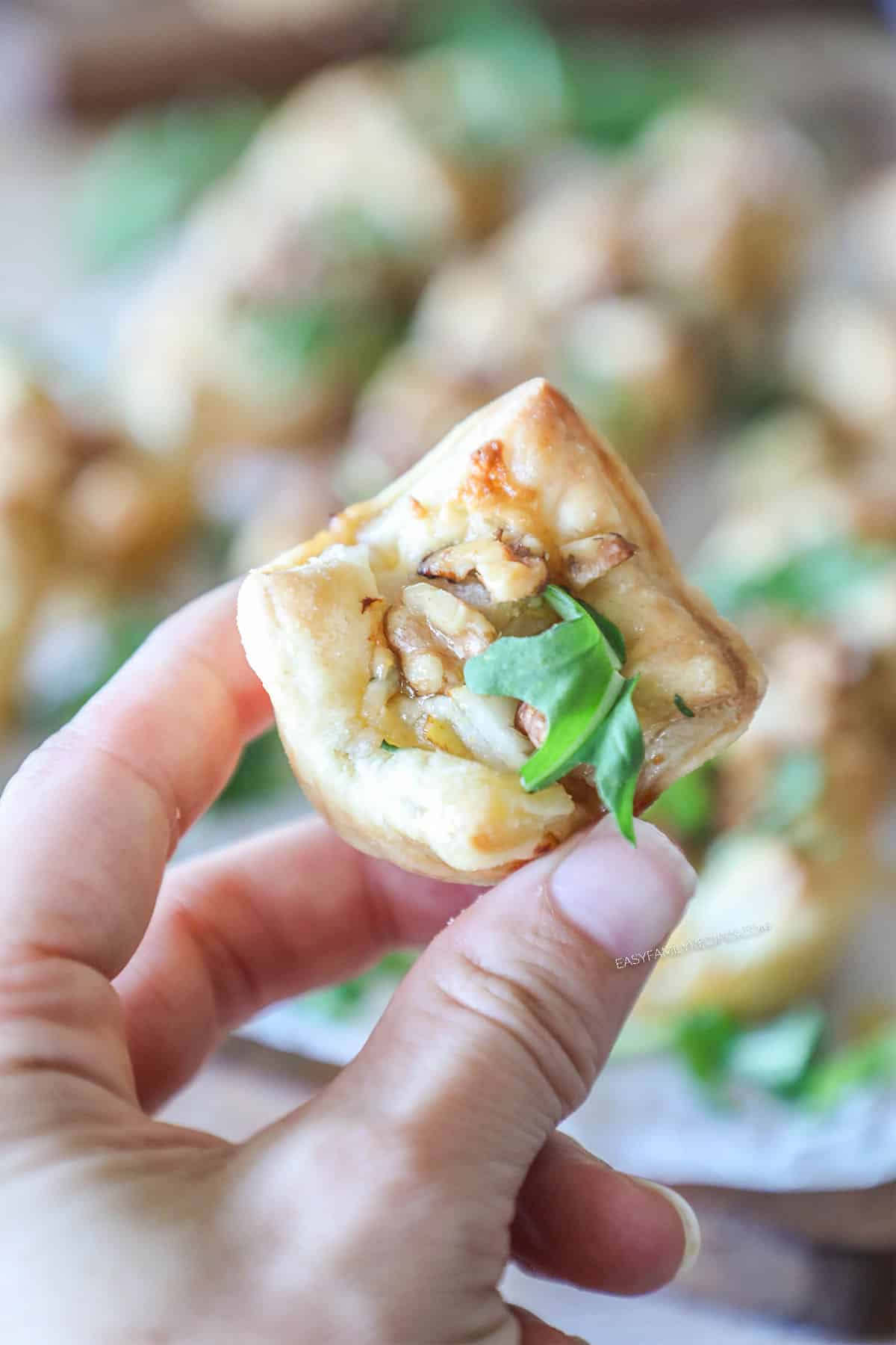 Close up of hand holding a single Pear and Gorgonzola Puff Pastry Bite. In the background are more bites on a wooden board
