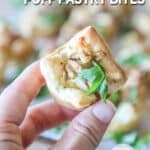 Close up of hand holding a single Pear and Gorgonzola Puff Pastry Bite. In the background are more bites on a wooden board