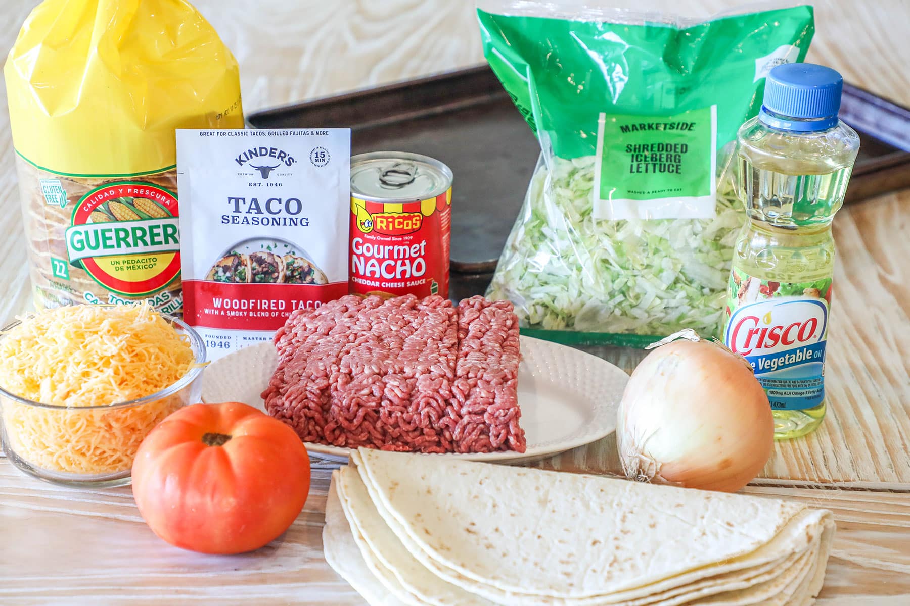 Ingredients for sheet pan crunch wrap supreme, including taco seasoning, nacho cheese sauce, lettuce, vegetable oil, corn tostadas, onion, tomato, ground beef, cheese, and flour tortillas.